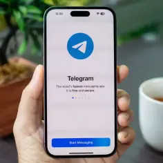 Dłoń trzyma smartfon z otwartą aplikacją Telegram, widoczne logo i przycisk "Start Messaging".