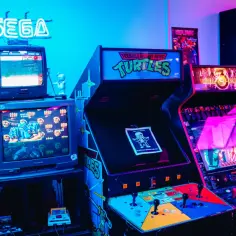 Neonowy napis SEGA nad starymi telewizorami i automatami do gier, gdzie królują gry retro: Teenage Mutant Hero Turtles i Mortal Kombat.
