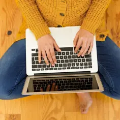 Kobieta w żółtym swetrze i jeansach pisze na laptopie, sprawdzając olx cennik ogłoszeń.