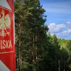 Słup graniczny z orłem i napisem Polska przy lesie. Dalej widać ogrodzenie z drutu kolczastego, symbolizujące strefę **dmz**.