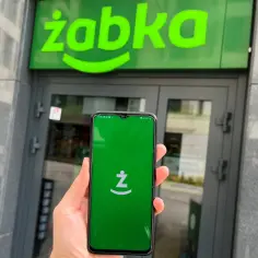 Dłoń trzyma smartfon z otwartą aplikacją Żappka przed wejściem do sklepu Żabka.