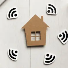 Dom z kartonu otoczony symbolami Wi-Fi, sugerujący sieć mesh. Obok fragmenty laptopa i routera.