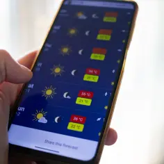 Dłoń trzyma smartfon z wyświetloną prognozą pogody. Widać ikony słońca i księżyca oraz temperatury, co czyni ją najlepszą aplikacją pogodową.
