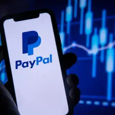Dłoń trzyma smartfon z logo PayPal. W tle wykresy finansowe, sugerujące jak płacić PayPal online.