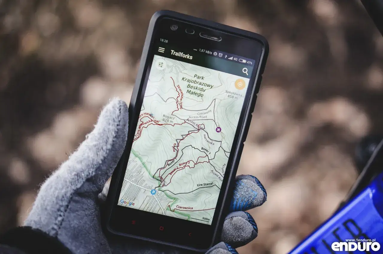 Ręka w rękawiczce trzyma smartfon z aplikacją na rower Trailforks, pokazującą mapę szlaków w Parku Krajobrazowym Beskidu Małego.