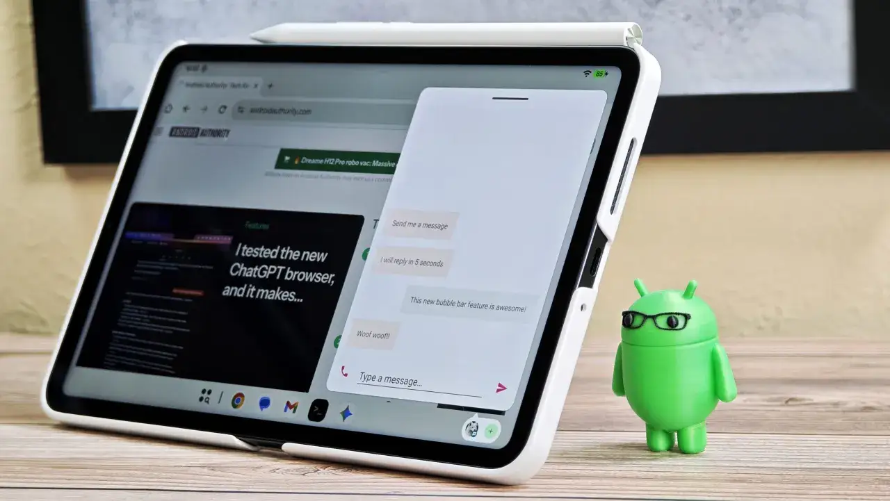 Zielony android 16 z okularami stoi obok tabletu z otwartą stroną Android Authority i aplikacją do czatowania.