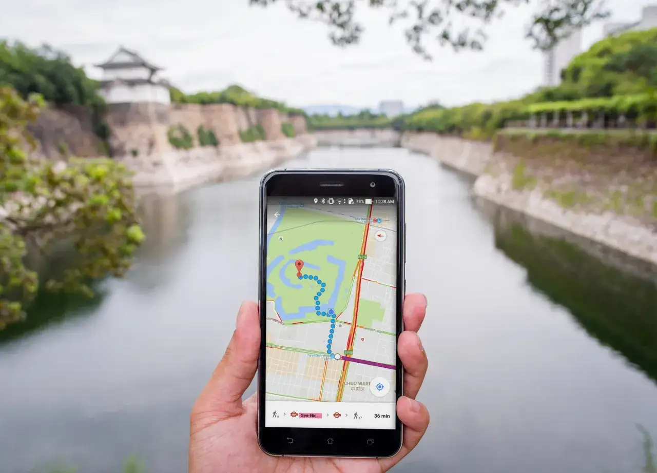 Dłoń trzyma smartfon z mapą Google, pokazującą trasę spaceru wokół zamku. Widok na fosę i mury obronne. google maps współrzędne pomagają odkrywać świat.