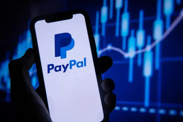 Dłoń trzyma smartfon z logo PayPal. W tle wykresy finansowe, sugerujące jak płacić PayPal online.